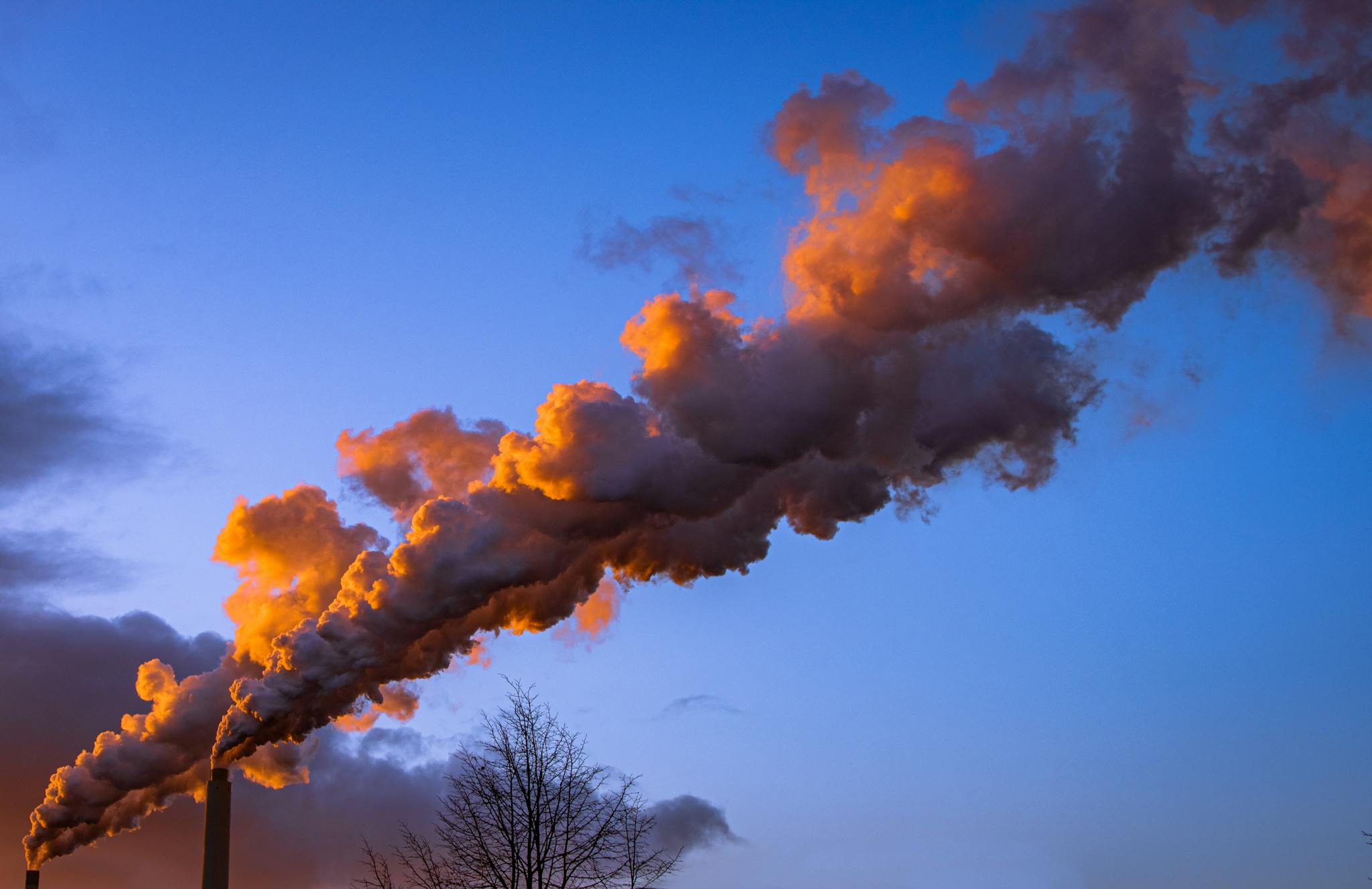 Industrial smokestack releasing orange-hued smoke during sunset against a vibrant sky.